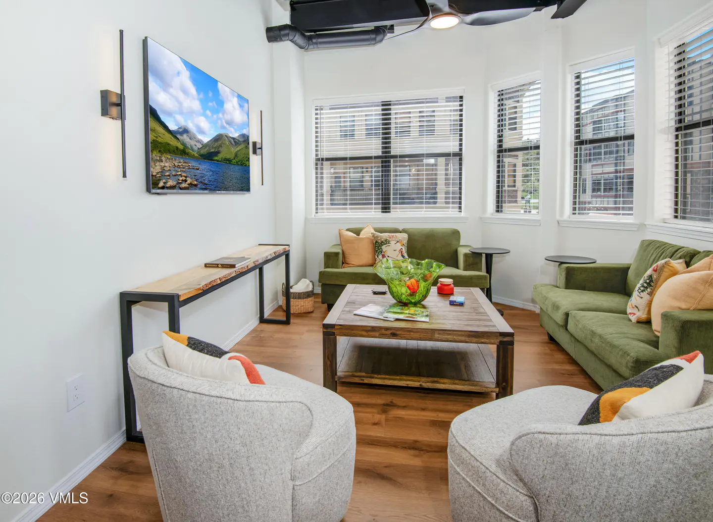 A bright living room with two green sofas, two gray chairs, and a wooden coffee table. A TV displays a mountain scene.