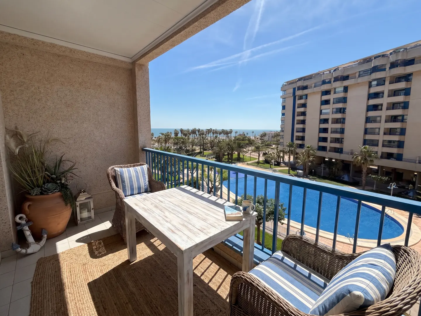Balcony with wicker chairs, a white table, and blue railings overlooking a pool, palm trees, and the ocean on a sunny day.