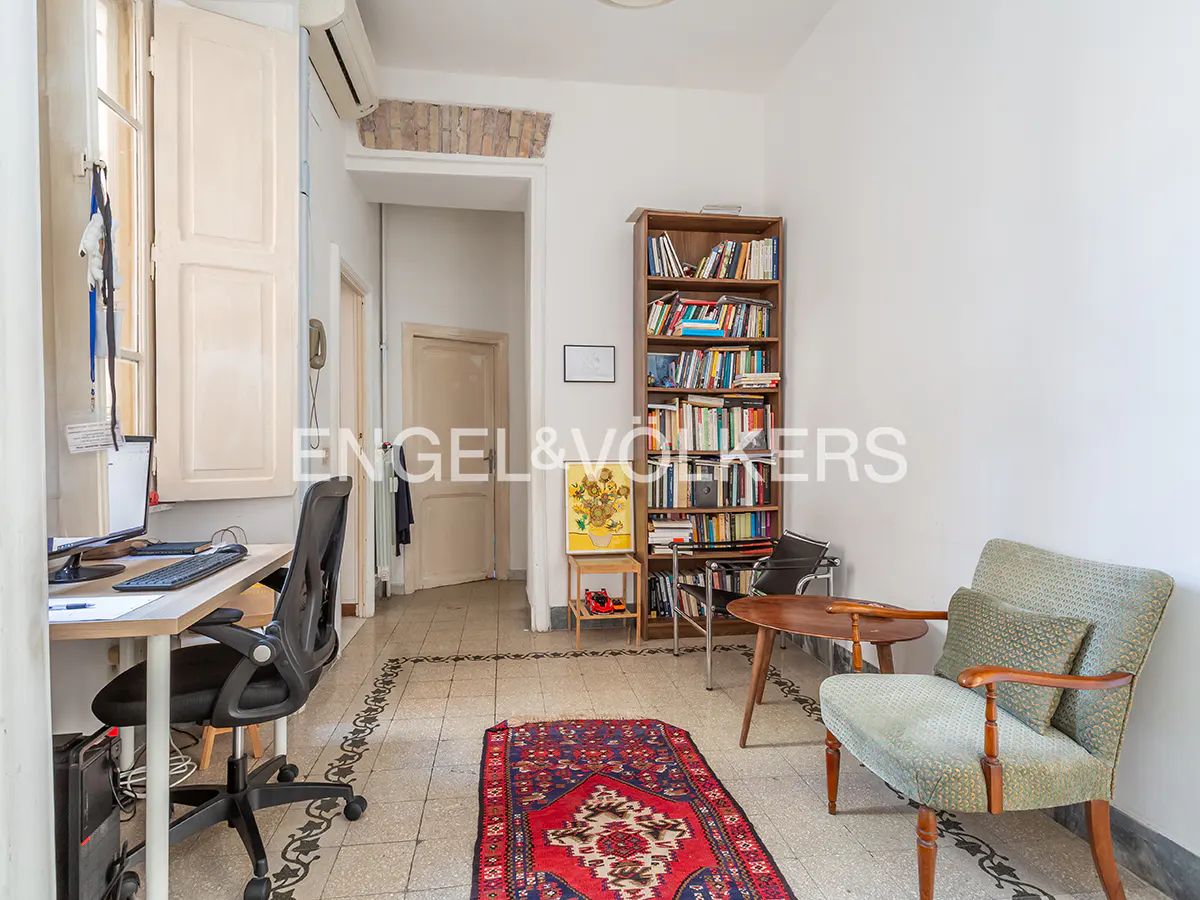 Home office with a desk, black chair, patterned rug, bookshelf, and a green armchair on a tiled floor.