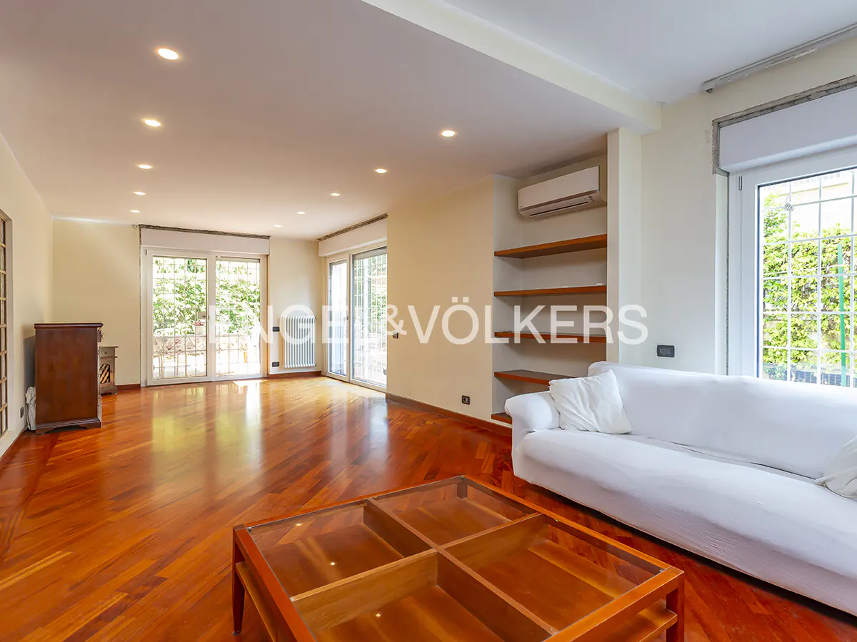 Bright living room with hardwood floors, white sofa, and glass coffee table. Sliding glass doors lead to a green outdoor space.