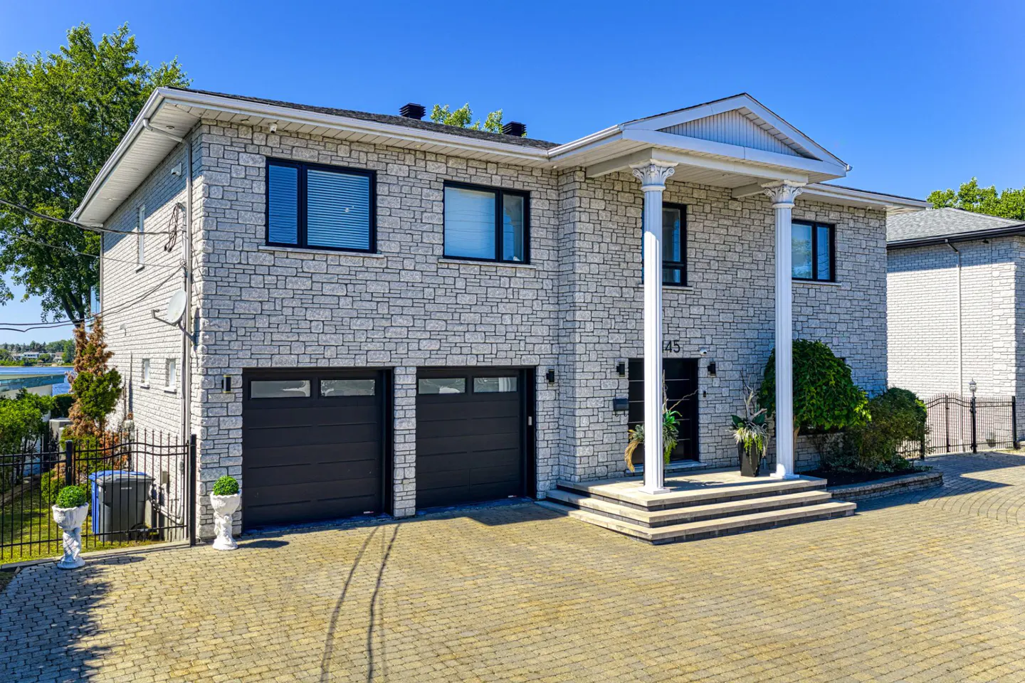 Two-story stone house with black garage doors and white columns at the entrance. Paved driveway and blue sky.