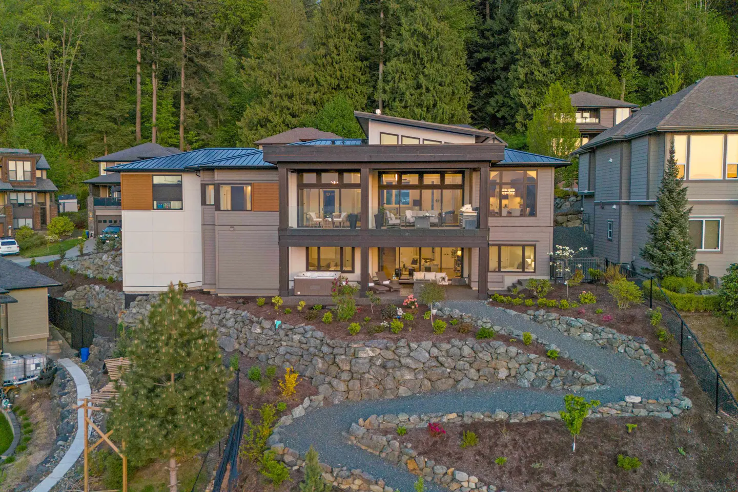 Two-story home with a gray exterior, large windows, and a balcony overlooking a stone-lined path and landscaped yard. Trees in background.