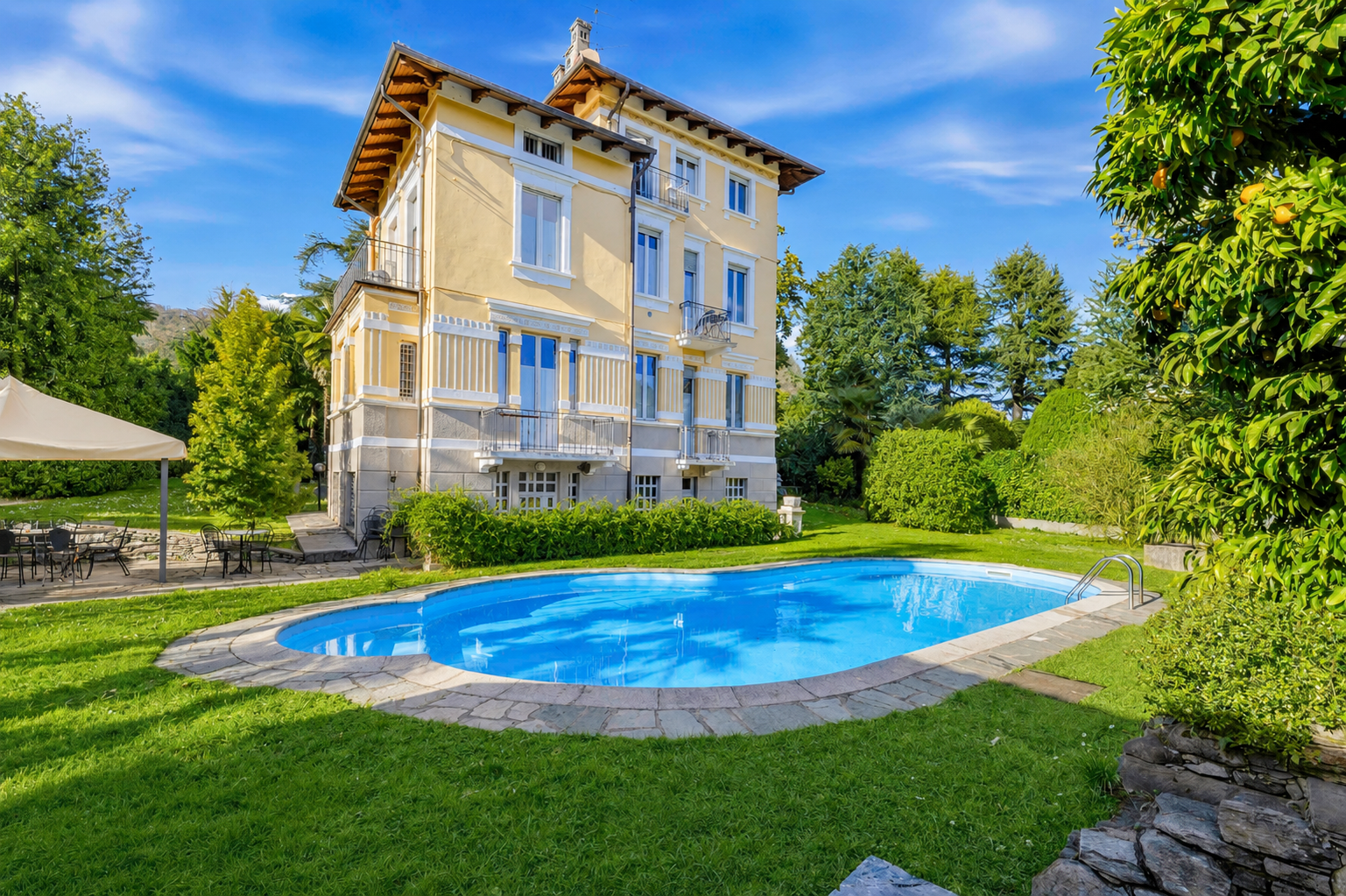 A yellow three-story villa with balconies overlooks a bright blue swimming pool in a lush, sunny garden.