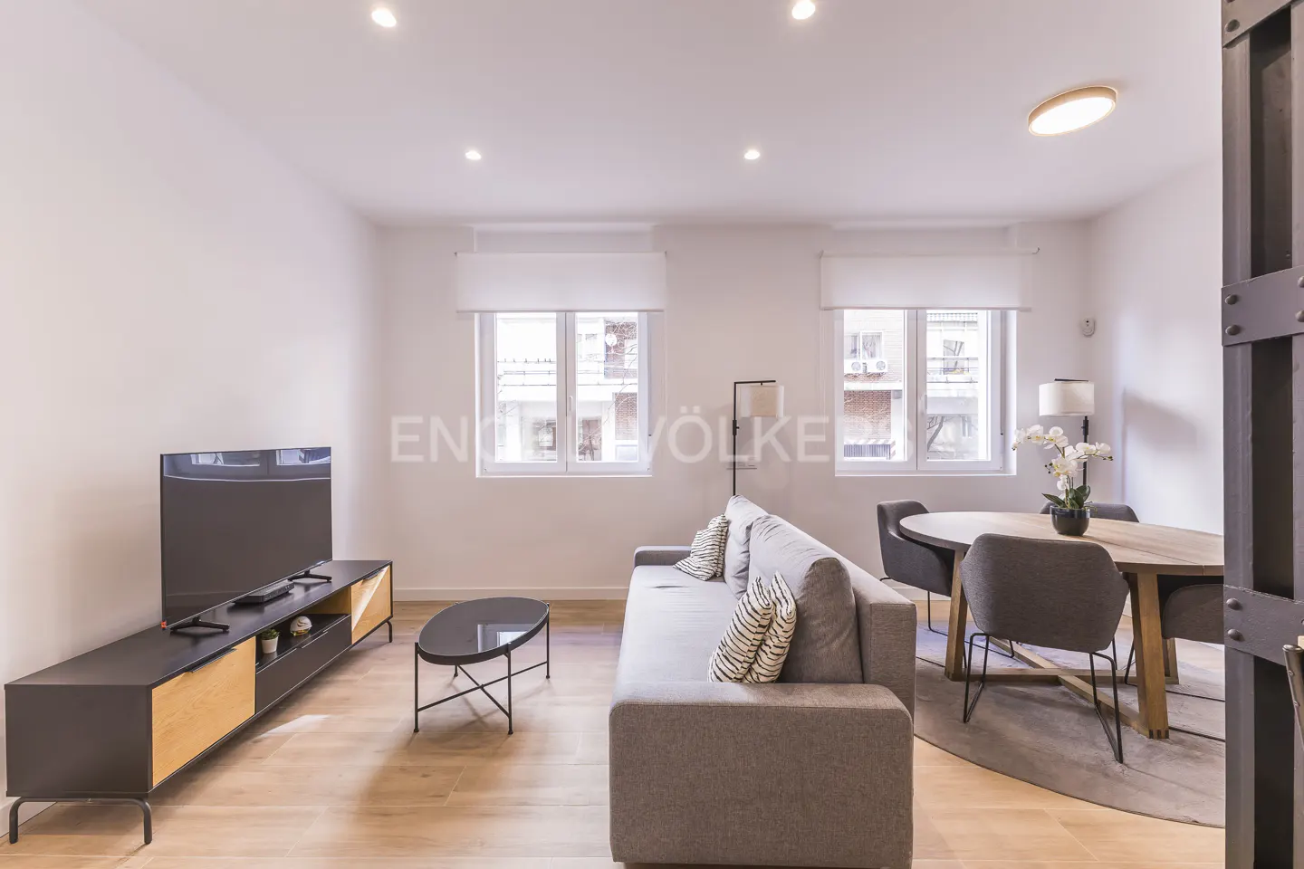 Modern living room with a grey sofa, oval coffee table, TV console, and a dining area with a wooden table and grey chairs.