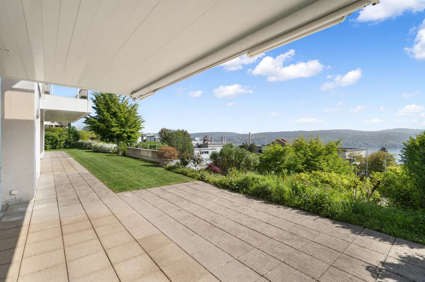 A patio with stone tiles overlooks a green lawn, trees, and a distant lake under a blue sky.
