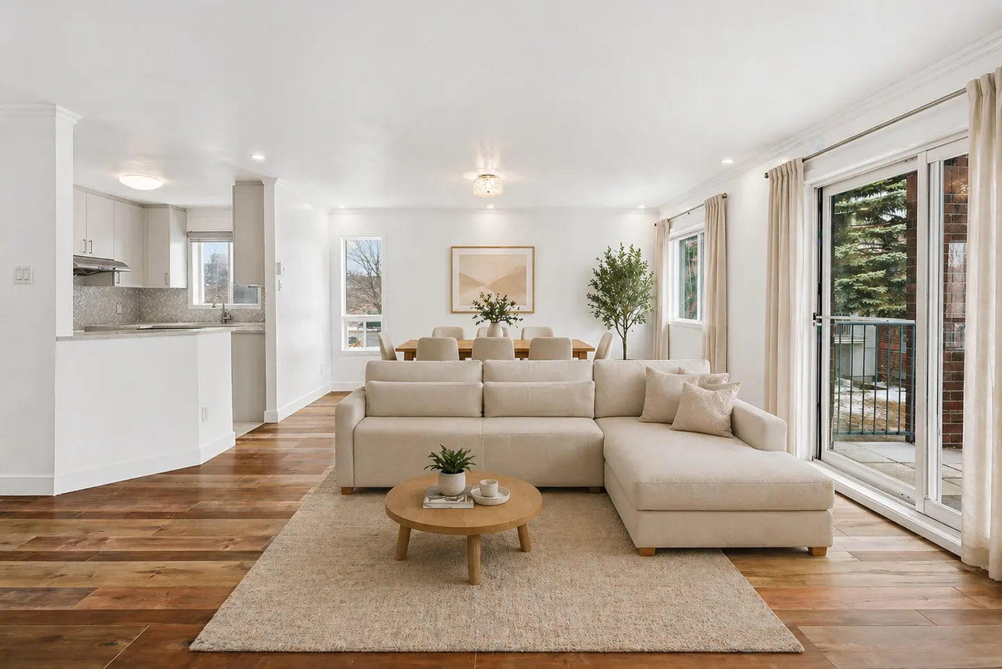 Bright living room with a beige sectional sofa, round coffee table, and dining area with a wooden table and chairs. Sliding glass doors lead to a balcony.