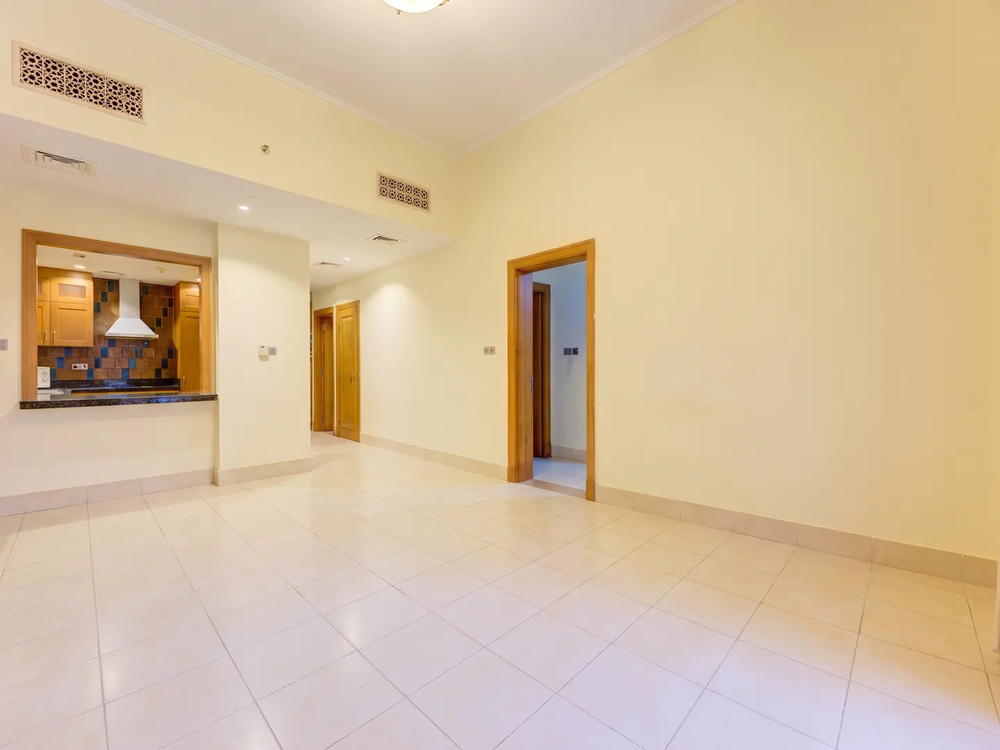 An empty room with cream walls and tiled floor, showing a kitchen with wooden cabinets and a white range hood.