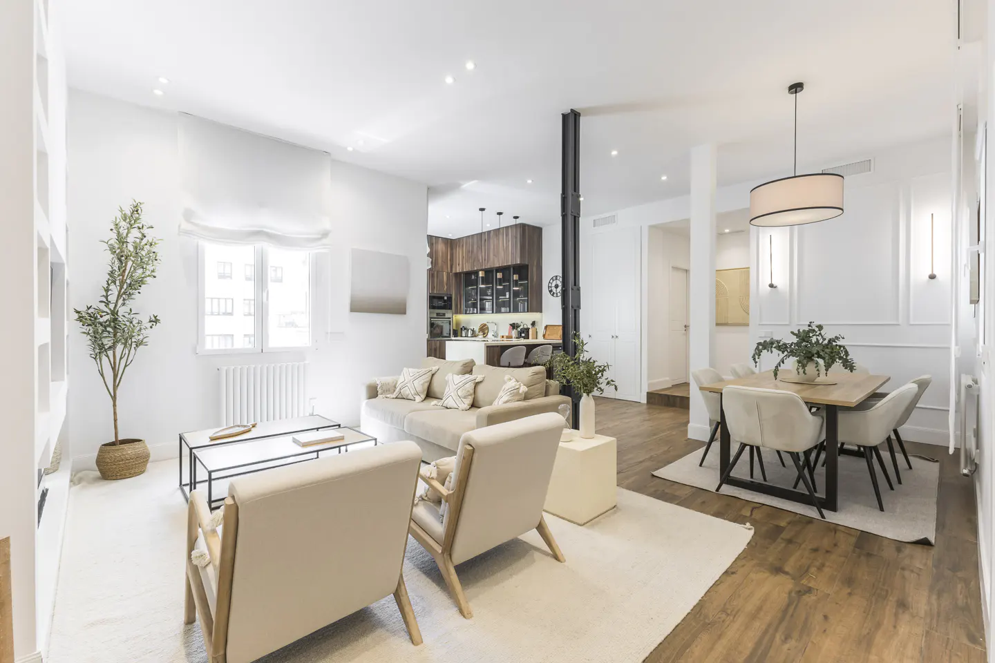 Bright, open-concept living space with a beige sofa, two armchairs, and a dining table with six chairs. A modern kitchen is visible in the background.