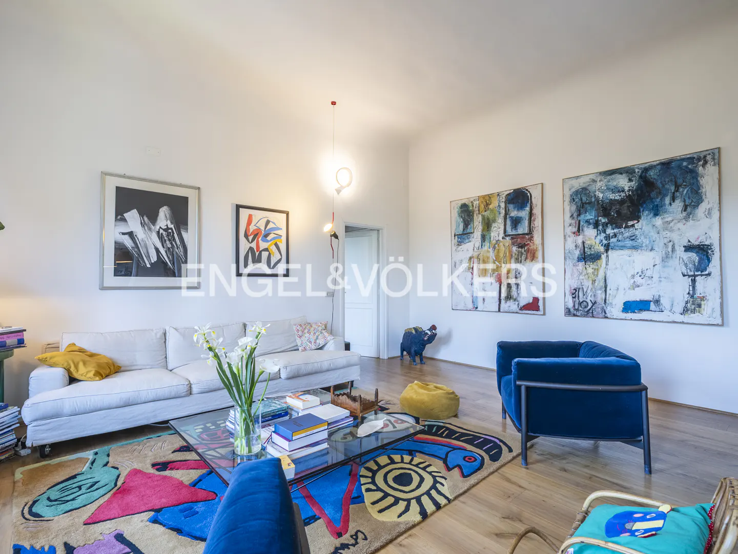 Bright living room with white walls, wood floors, and colorful rug. A white sofa, blue chairs, and abstract art add to the modern design.