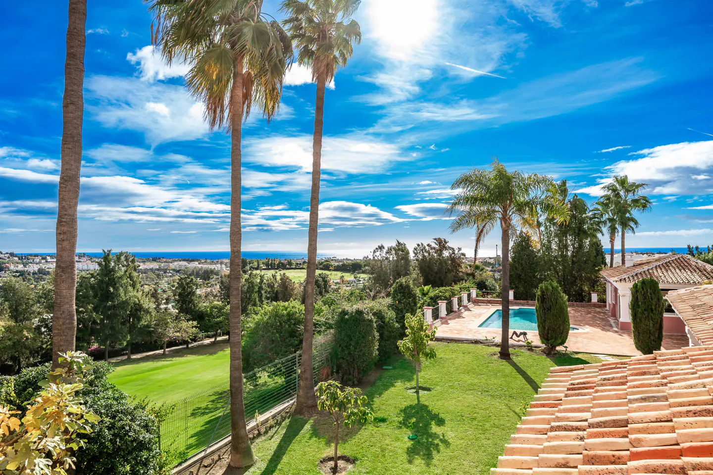 Scenic view of a property with a pool, palm trees, and a red tile roof under a blue sky with scattered clouds.