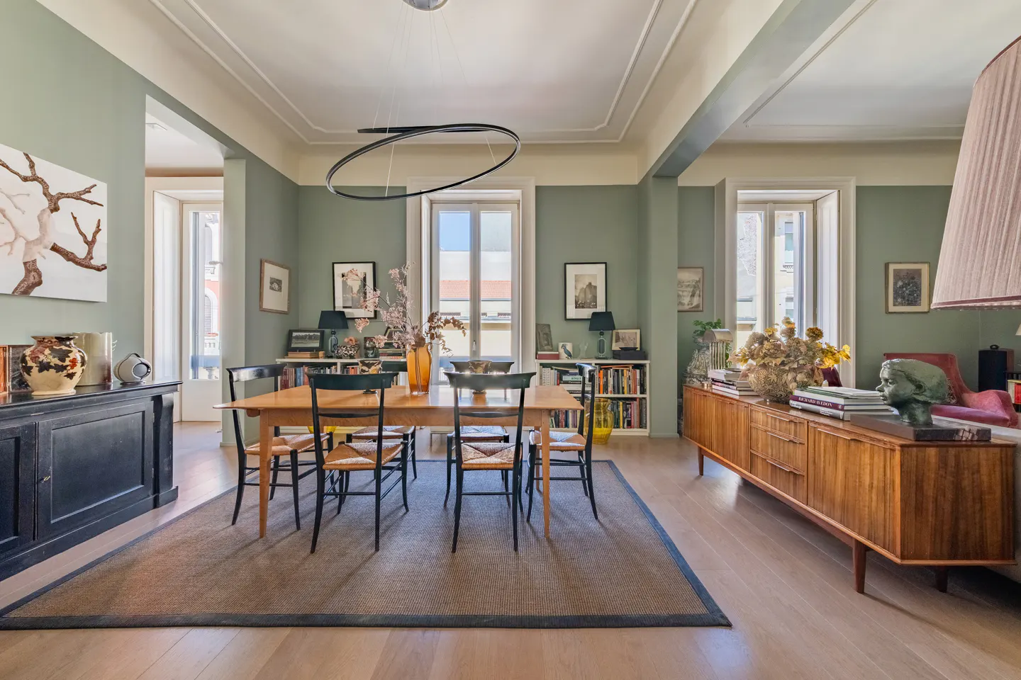 A dining room with sage green walls, a wooden table with black chairs, and a brown rug. A black sideboard and a wooden cabinet are on either side of the room.