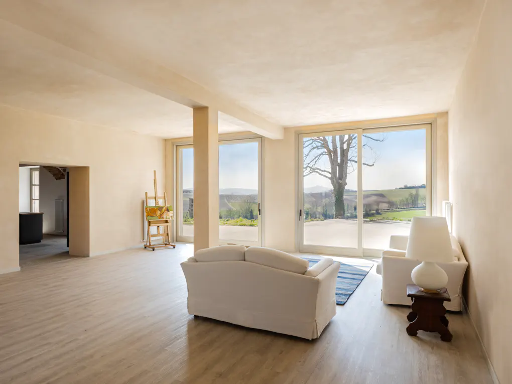 Bright, airy living room with white sofa, chair, and large windows overlooking a green landscape. An easel stands near the windows.