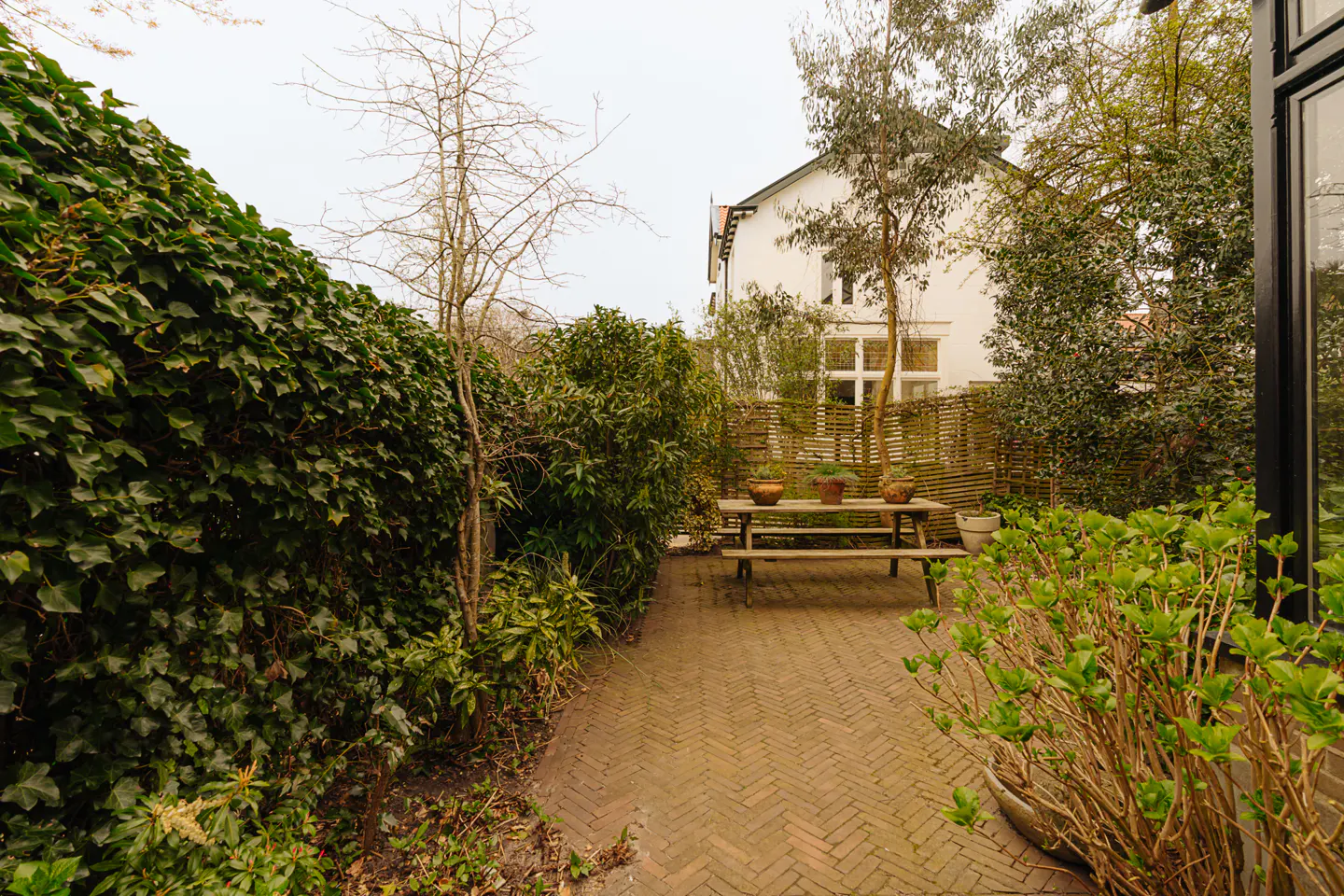 A brick patio with a picnic table and potted plants, surrounded by green hedges and a white house in the background.