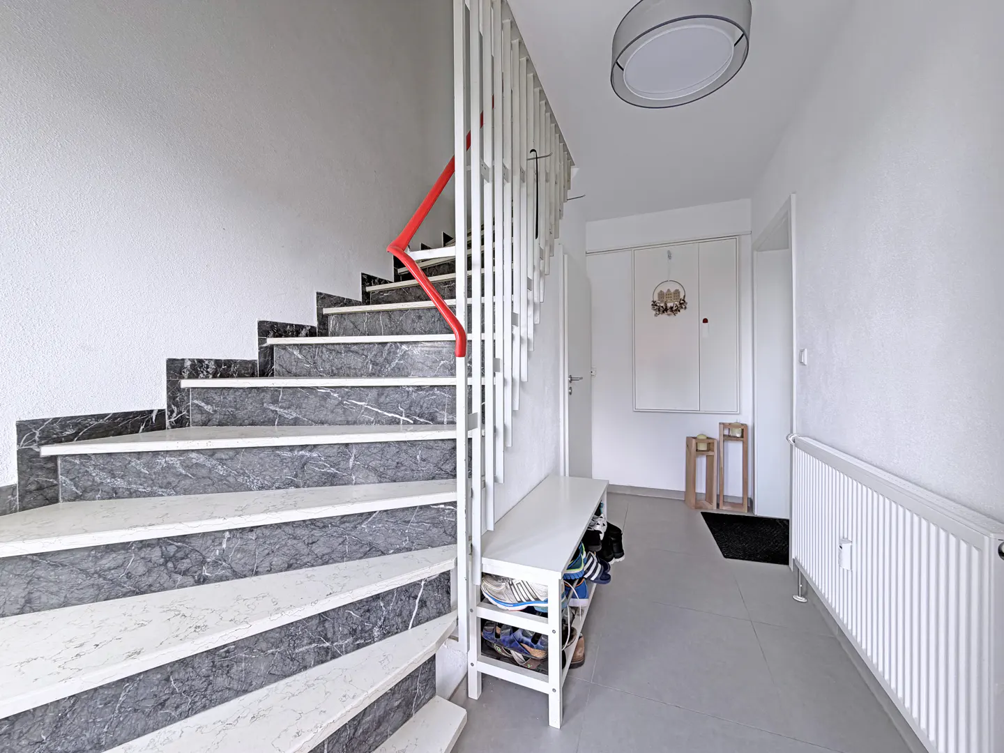 Staircase with grey marble steps and a white railing with a red handrail, leading to a bright, modern entryway.