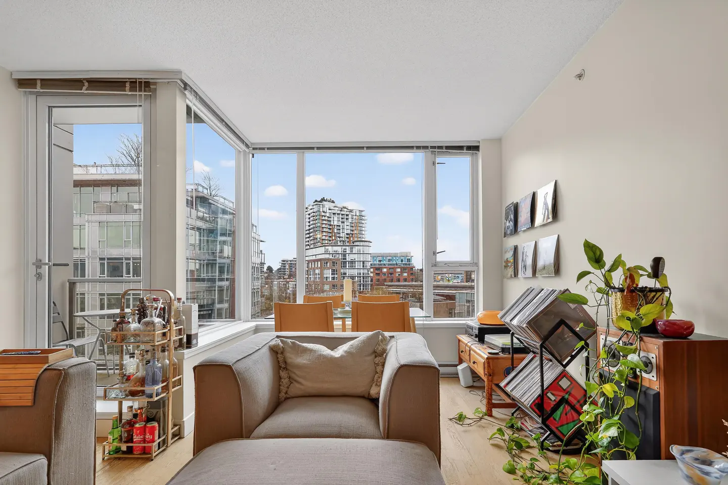 Bright living room with a gray armchair, floor-to-ceiling windows showing a city view, and a bar cart. Vinyl records and plants add character.