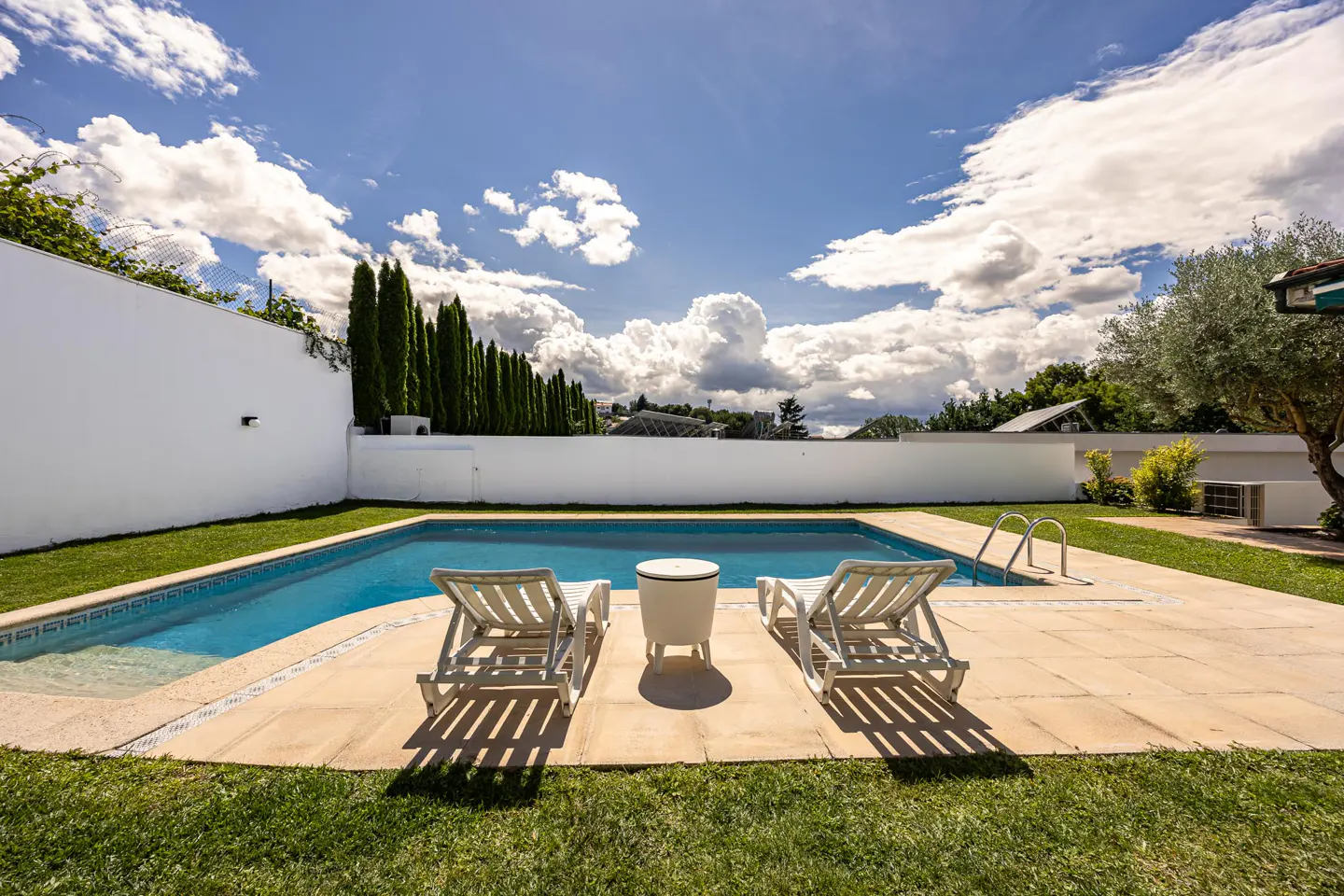Outdoor pool area with two white lounge chairs, a small table, and a blue pool surrounded by green grass and a white wall.
