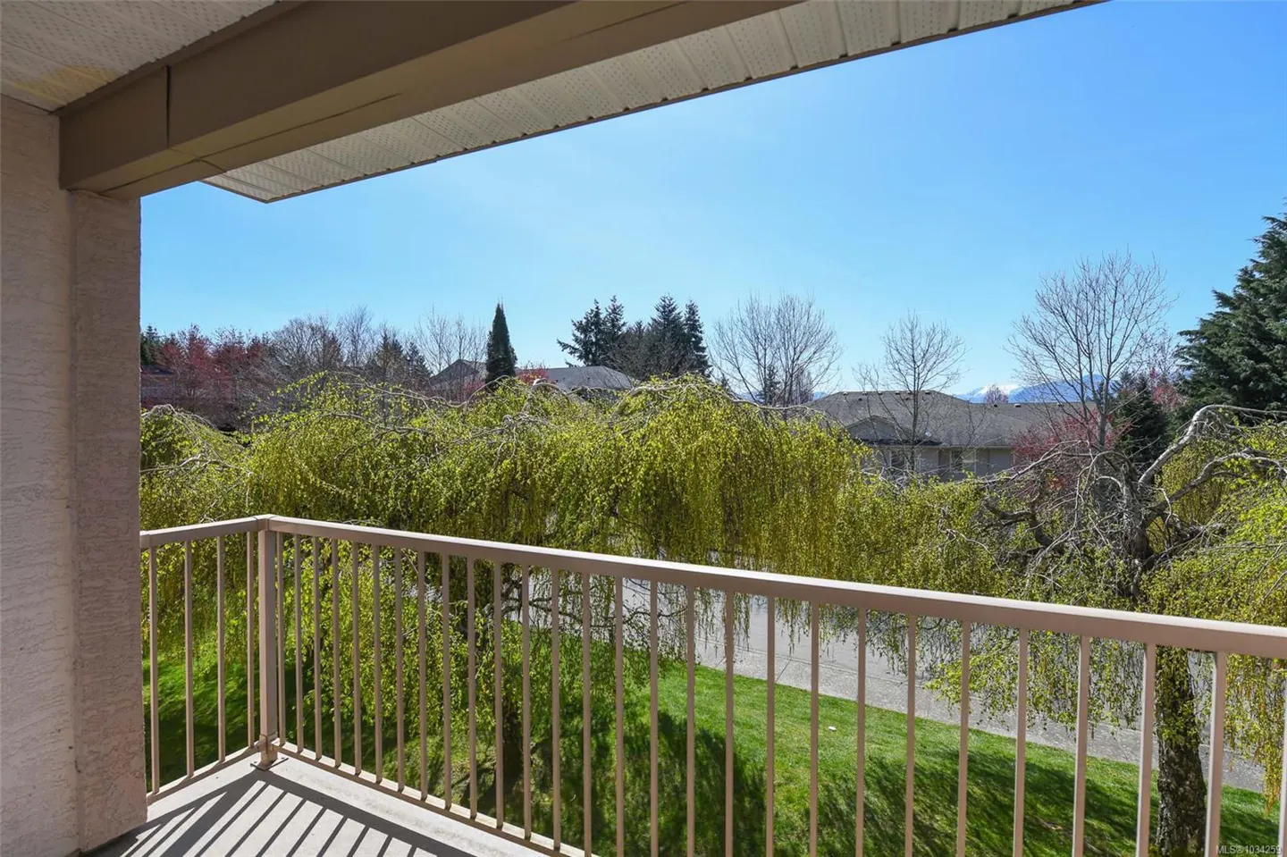 Balcony view with beige railing overlooking green trees, grass, and houses under a clear blue sky.