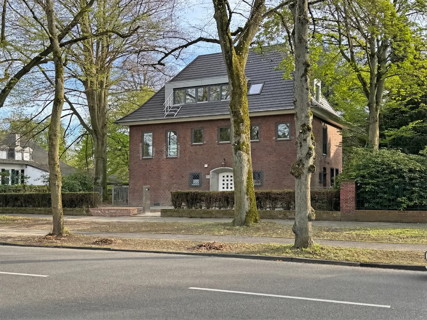 A two-story brick house with a gray roof is surrounded by trees and greenery. A white door is centered on the front facade.