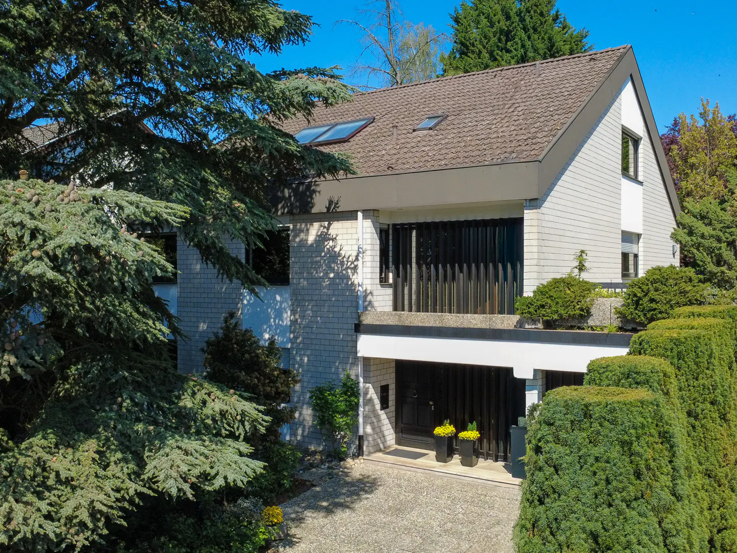Two-story house with gray shingles, brown roof, and black garage door. Green trees surround the house.