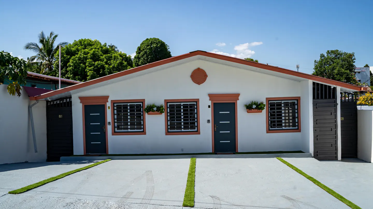 A white, one-story house with a brown trim, two dark gray doors, and two windows. There is a gray driveway with strips of artificial grass.