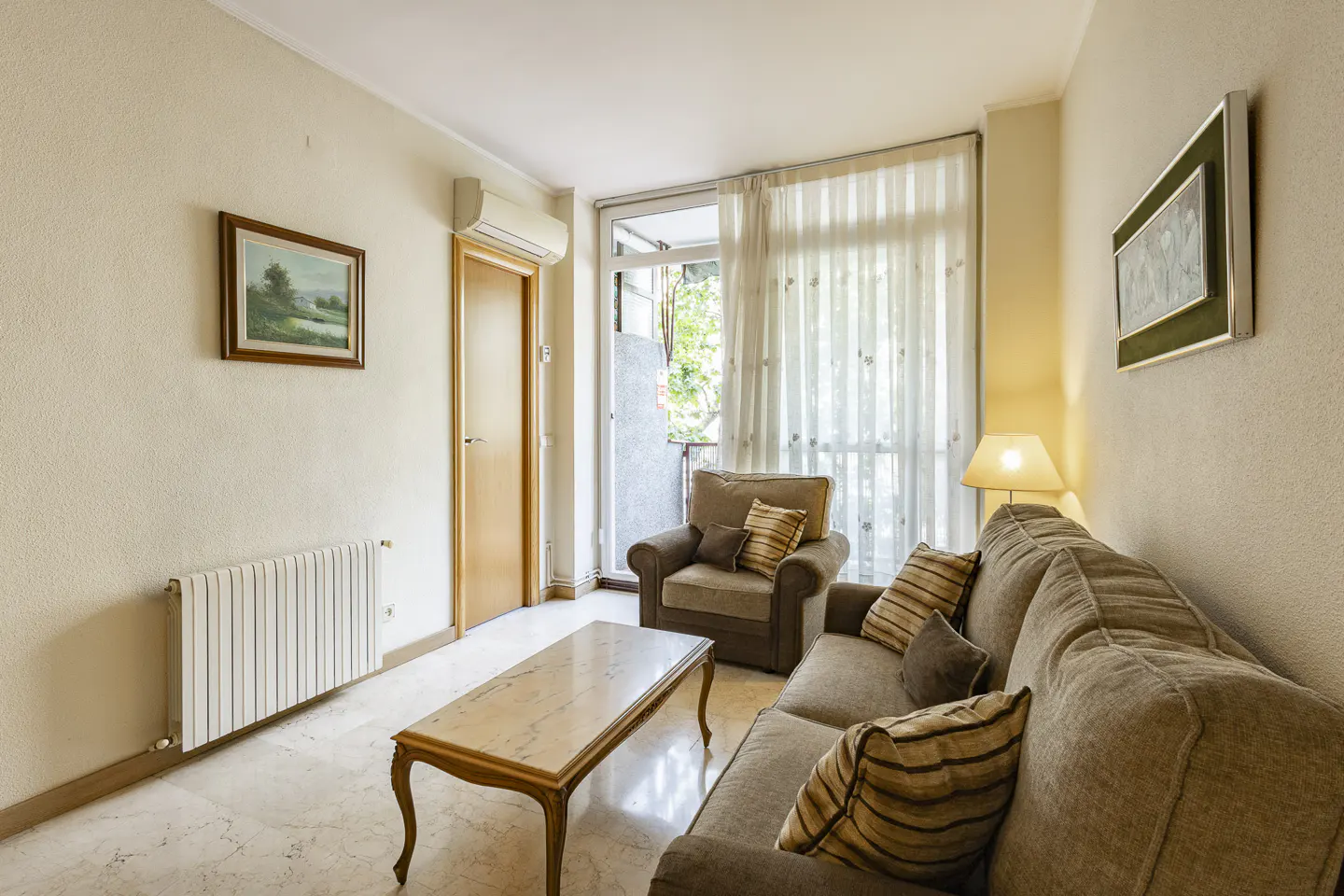 Living room with beige sofa, armchair, marble table, and balcony with sheer curtains. Artwork hangs on the walls.