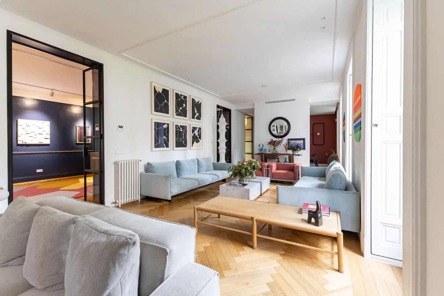 Bright living room with light blue sofas, wood floors, and white walls adorned with black and white art. A black framed glass door leads to a colorful room.