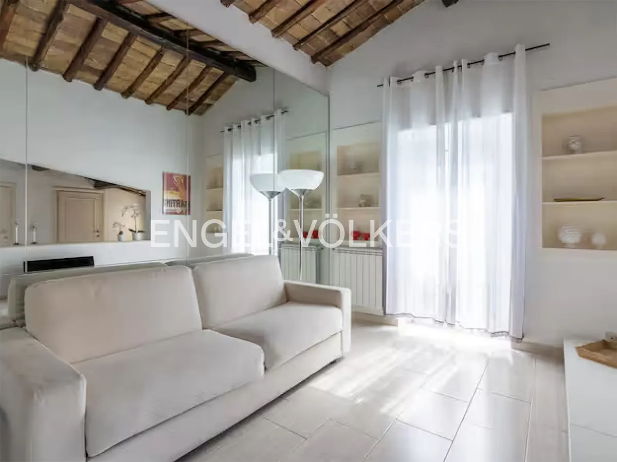 Bright living room with white sofa, tile floor, and exposed beam ceiling. Large mirror reflects the room. White curtains filter sunlight.