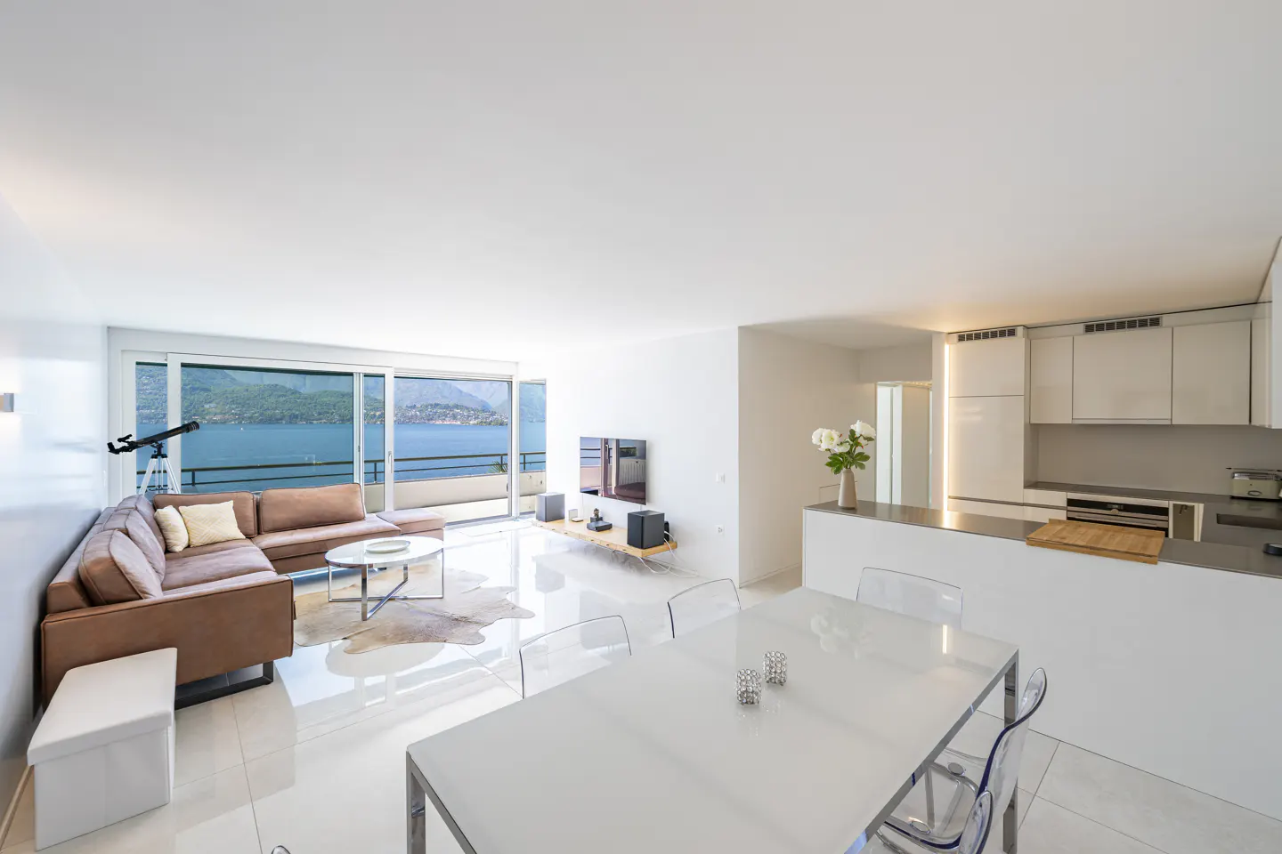 Bright, modern apartment interior with a brown sectional sofa, white dining table, and a view of a blue lake. A telescope stands near the sliding glass doors.
