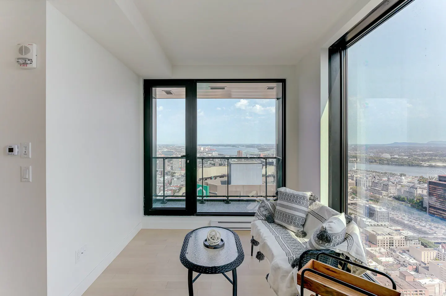 Bright sunroom with a black wicker table, a loveseat with patterned pillows, and a city view through large windows.
