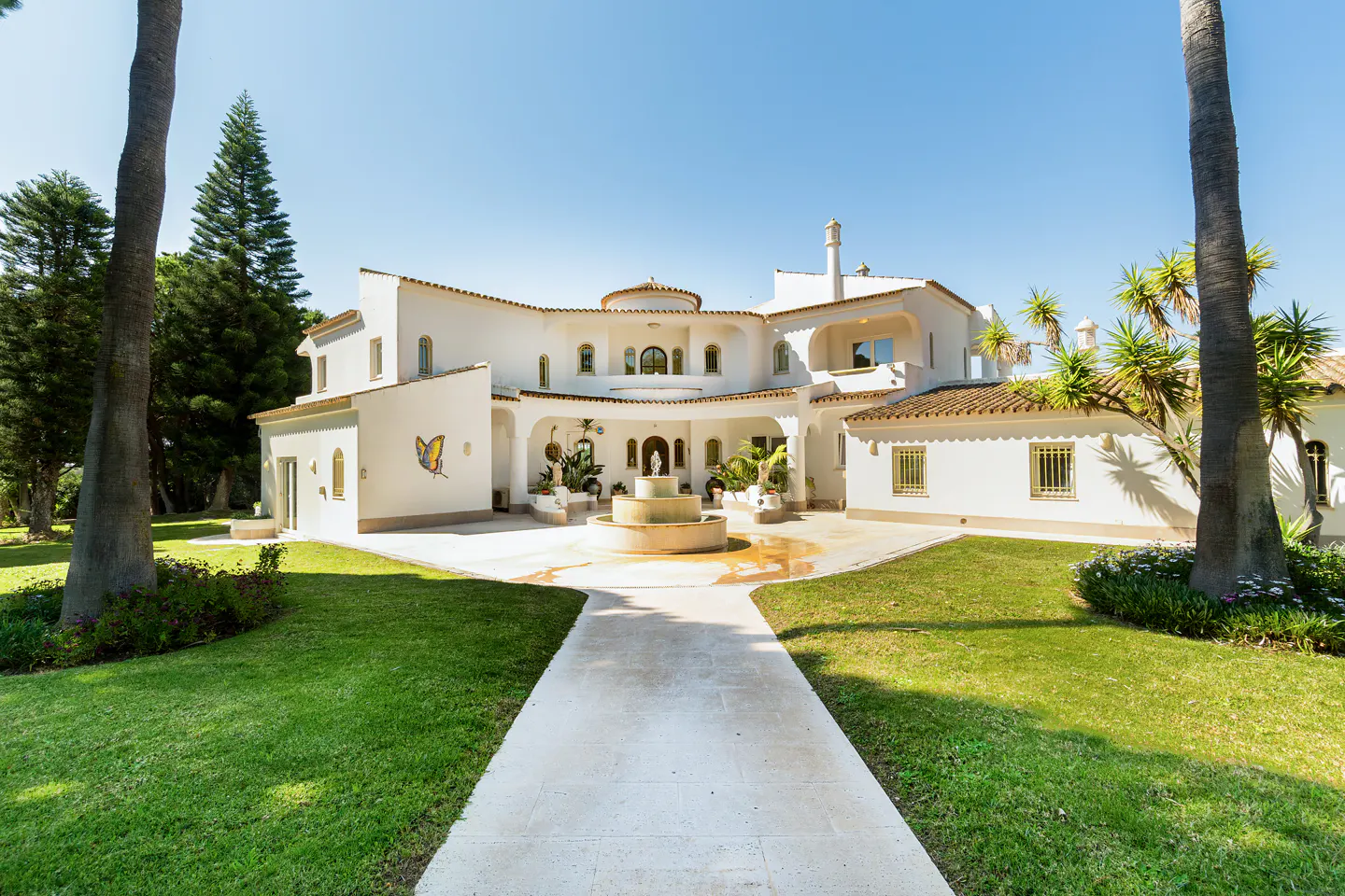 Exterior view of a large white house with a fountain, green lawn, and palm trees under a clear blue sky.