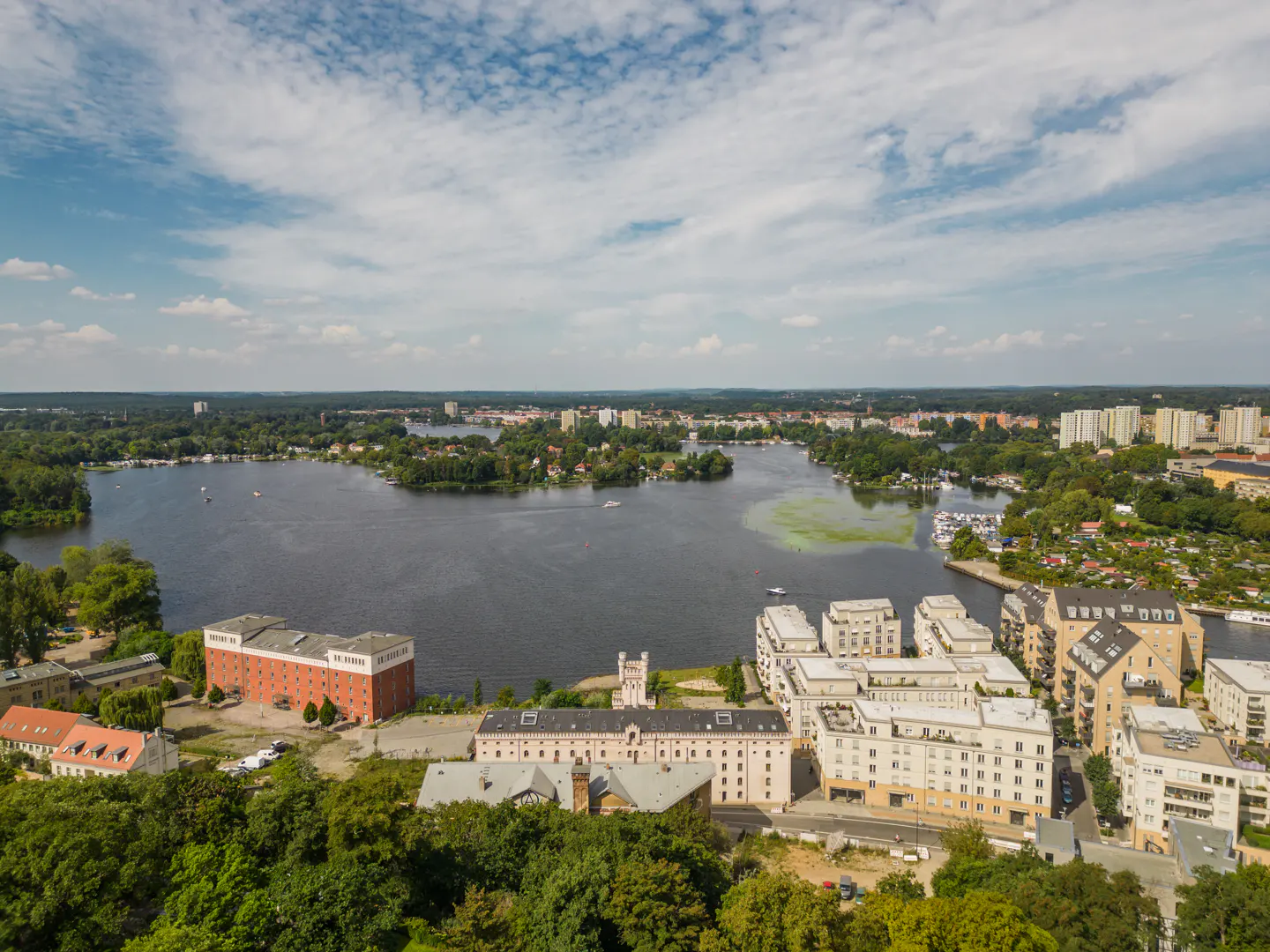 Aerial view of a lake surrounded by buildings and trees under a cloudy blue sky.