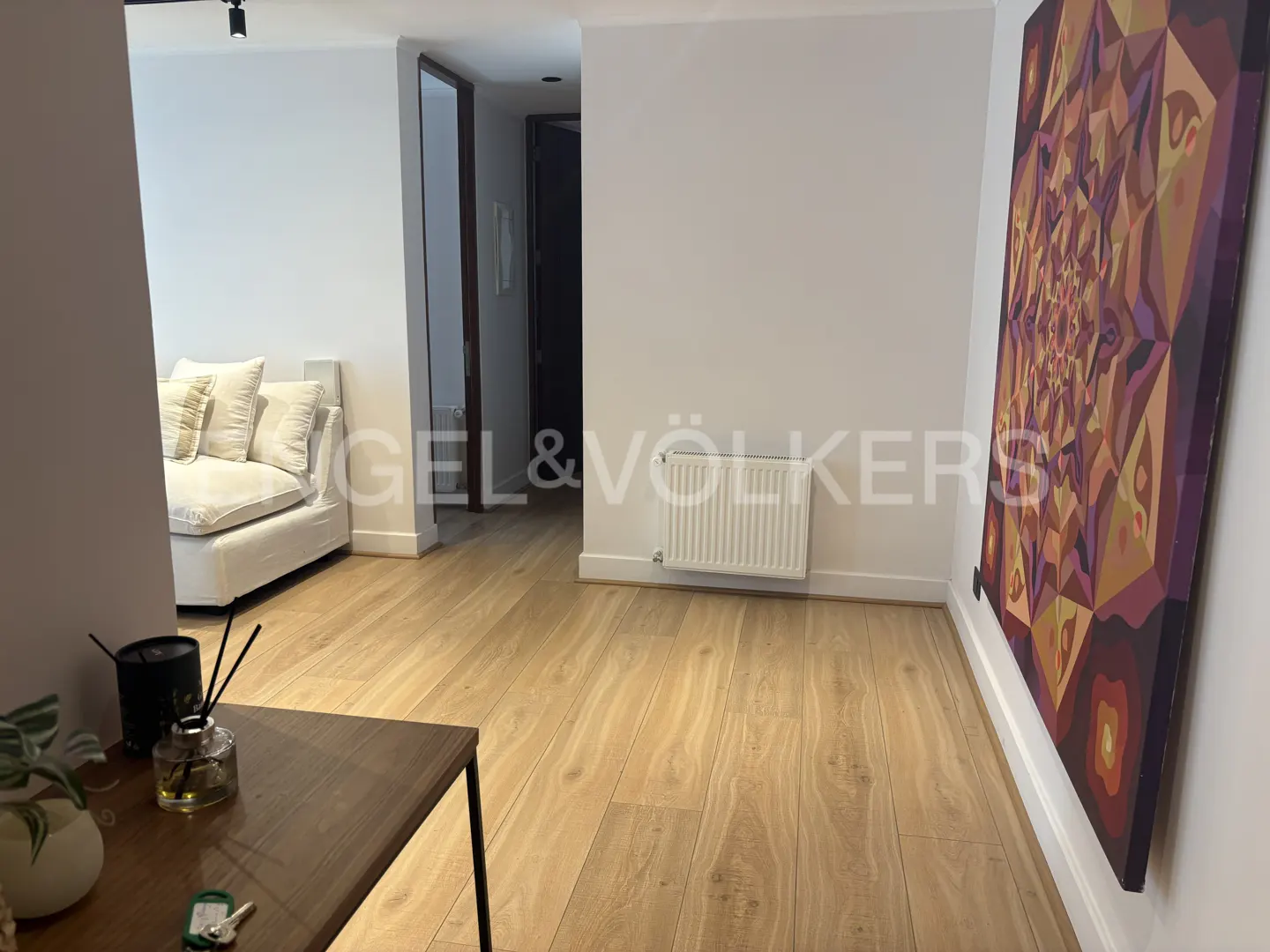 A bright living room with light wood floors, a white sofa, and a large, colorful abstract painting on the wall. A dark wood table is in the foreground.
