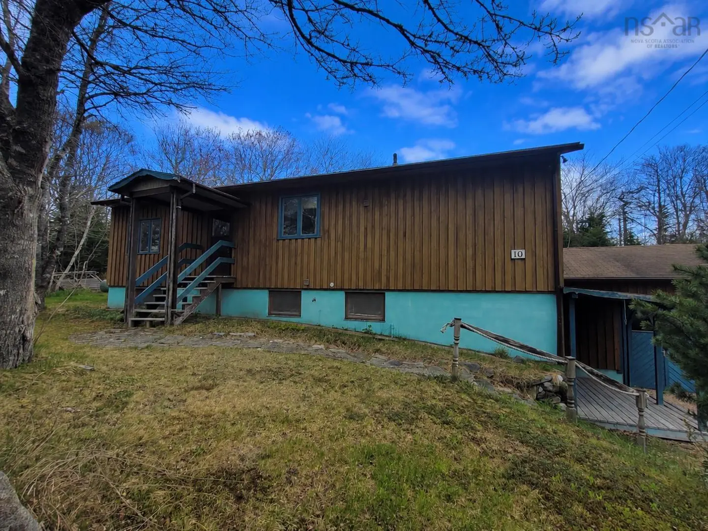 Exterior view of a two-story house with brown wood siding and a blue foundation, stairs, and window trim. The house has a porch and is surrounded by grass and trees.