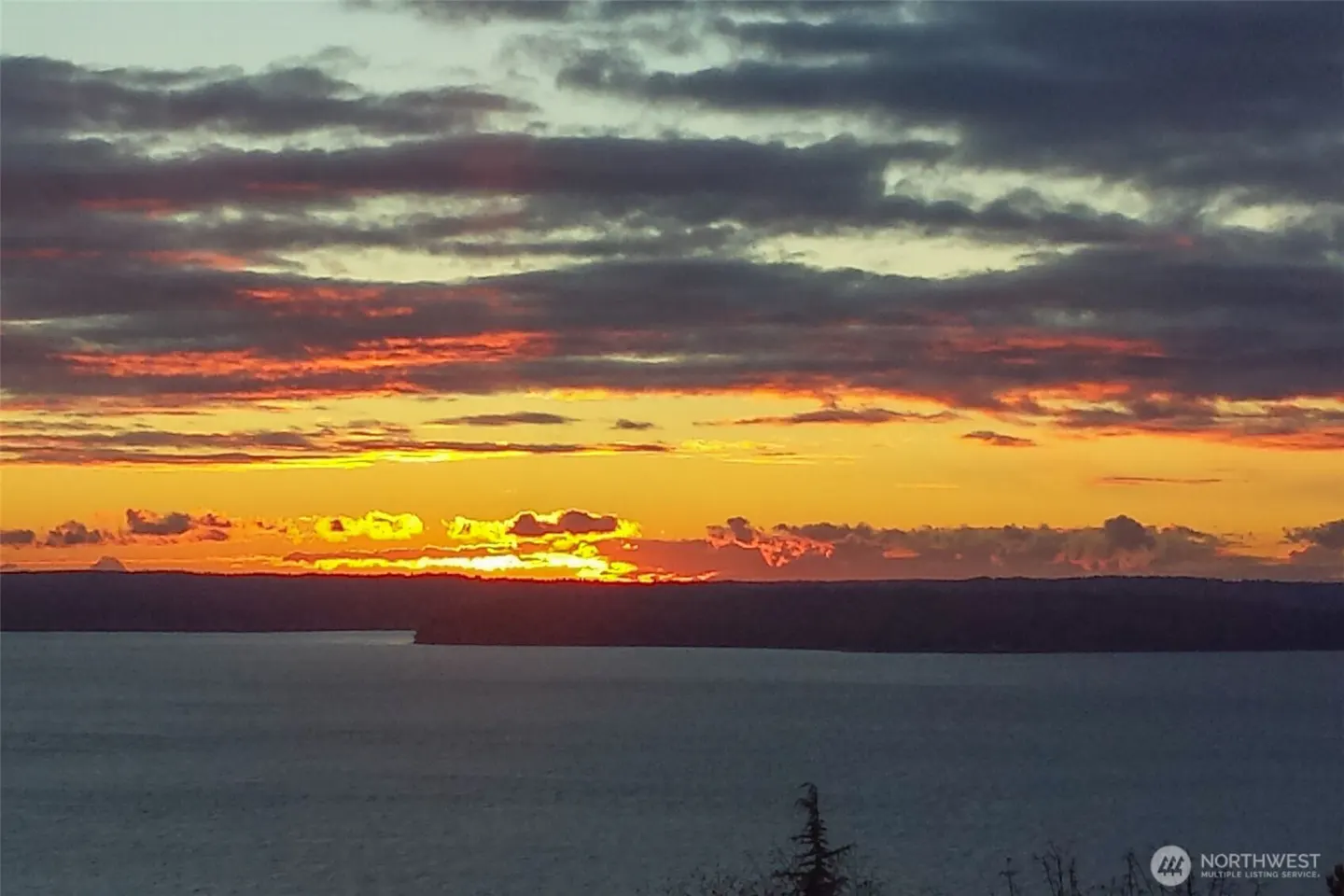 Sunset over water, with orange and gray clouds. Silhouetted land in the distance.
