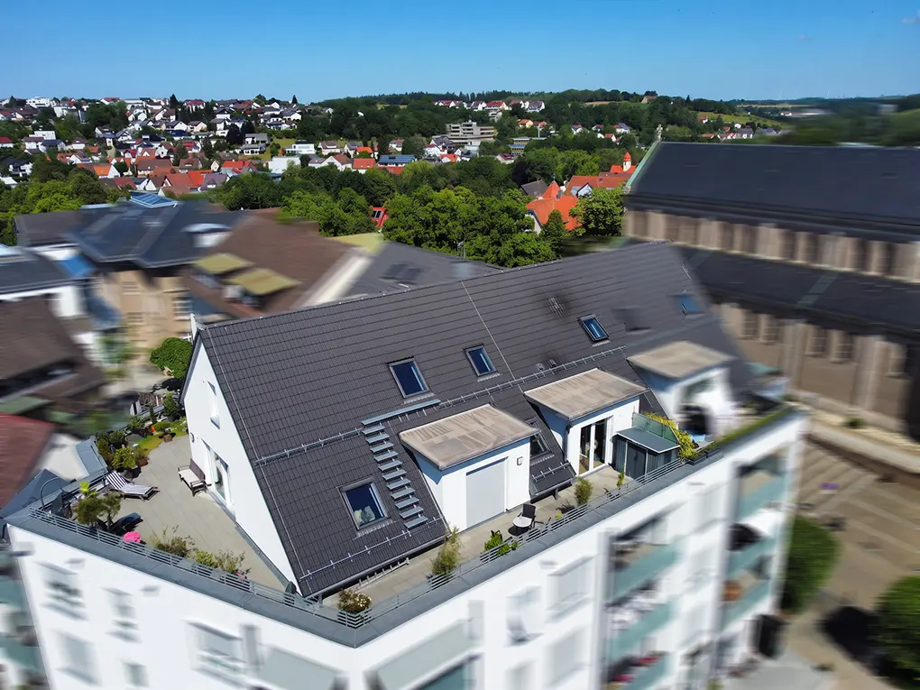 Aerial view of a white apartment building with a dark grey roof, rooftop terraces, and a surrounding town landscape.