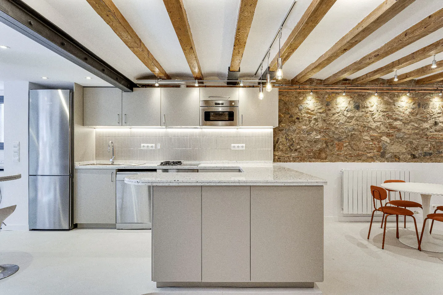 Modern kitchen with gray cabinets, stainless steel appliances, and a large island. Exposed wooden beams and a stone wall add rustic charm.