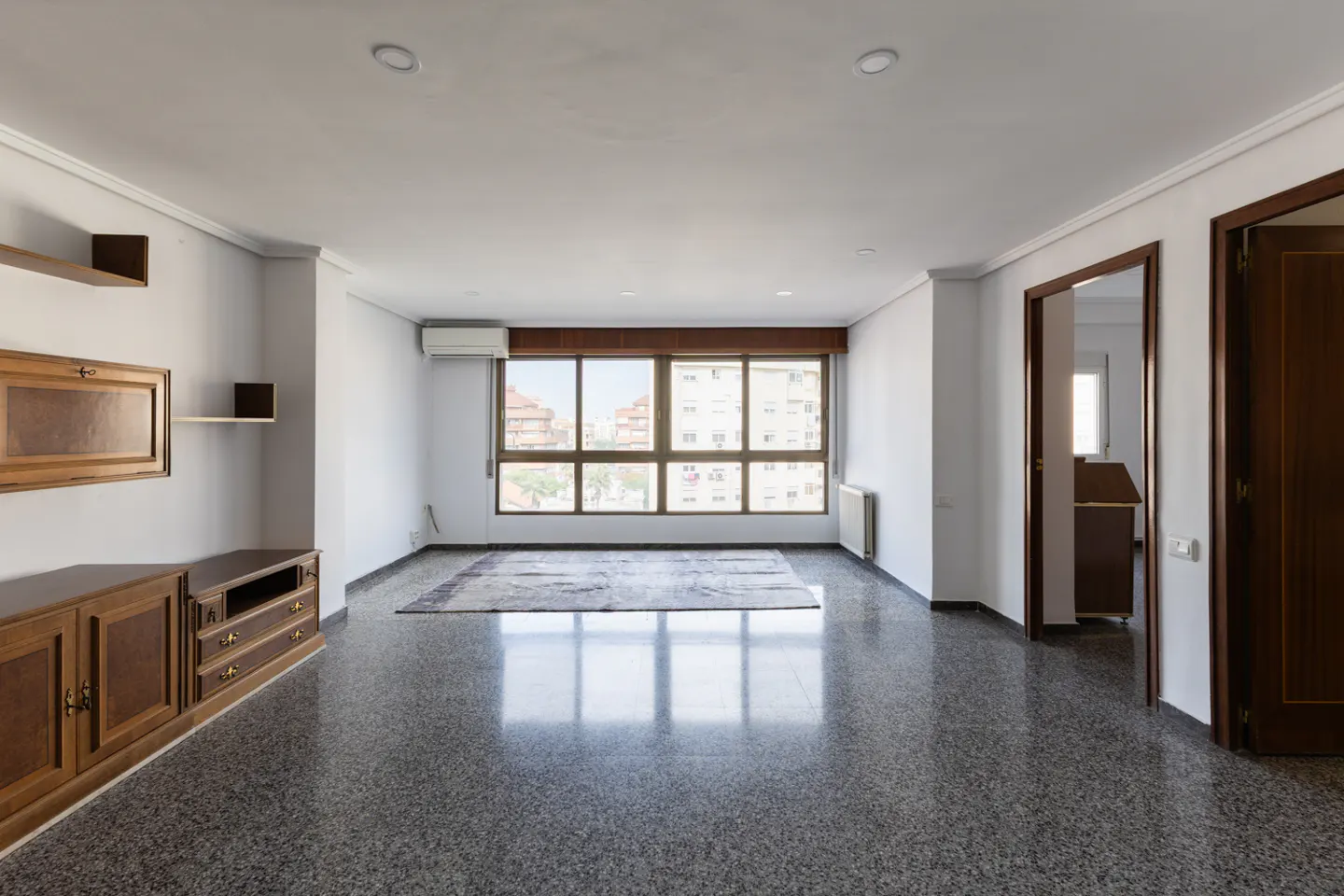 Empty living room with gray speckled floors, white walls, and a large window. A brown wooden cabinet is on the left wall.