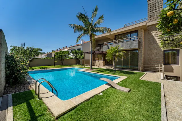 A modern stone house with a blue swimming pool, diving board, and palm trees on a sunny day.