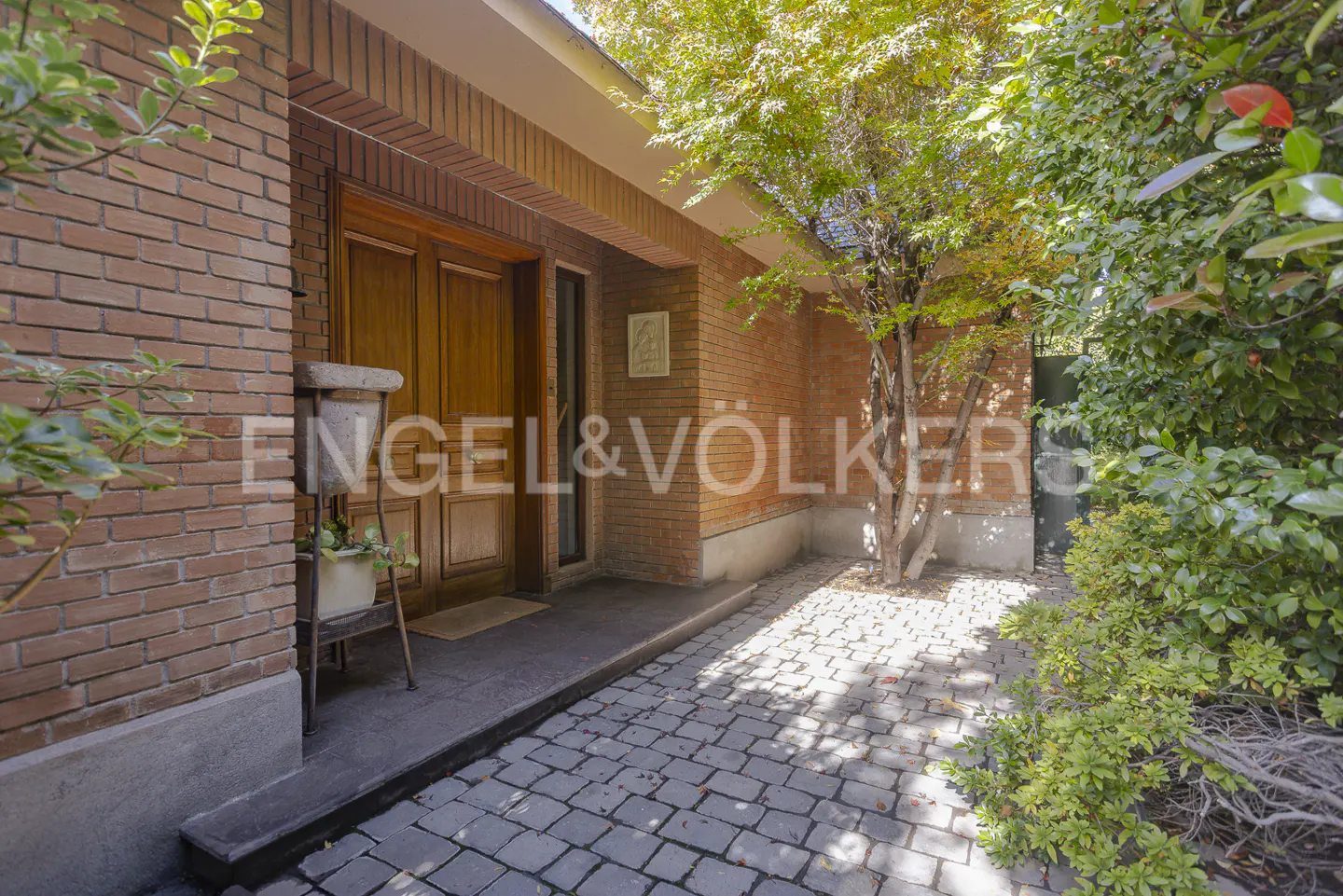 Brick house entrance with wooden door, stone walkway, and green foliage. Engel & Völkers logo visible on the brick wall.