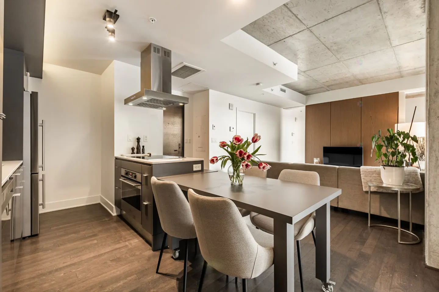Open-concept condo with a gray dining table, beige chairs, and a vase of red tulips. The kitchen has stainless steel appliances.