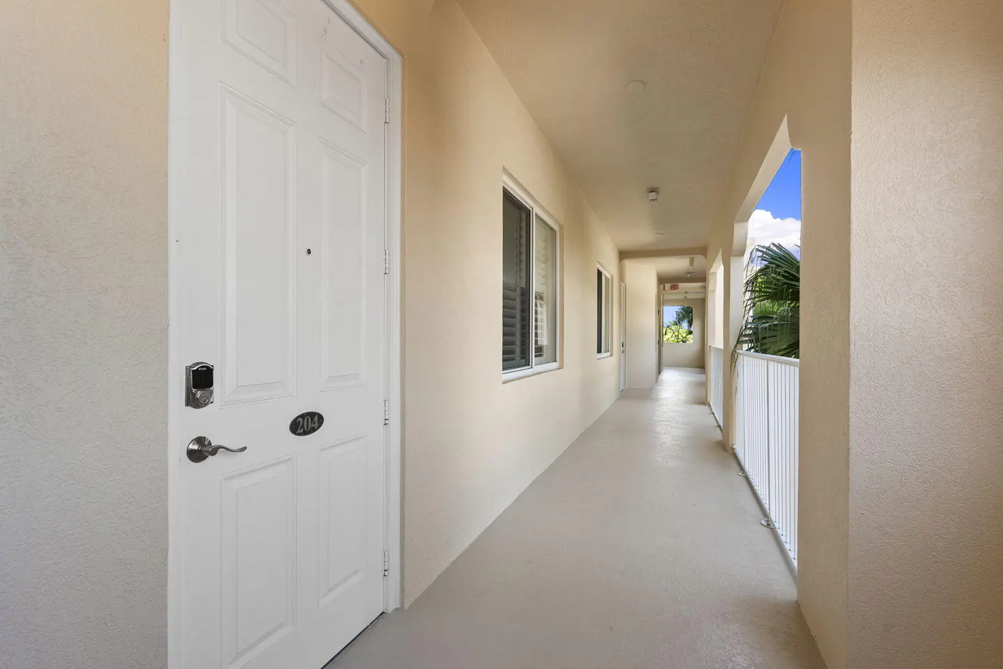 Exterior hallway of a condo building with a white door labeled 204, beige walls, and a white railing overlooking palm trees.