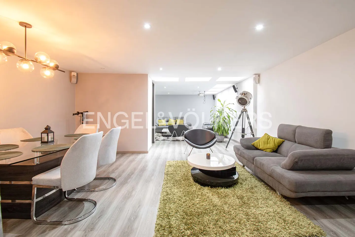 A modern living room with a gray sofa, green rug, and dining table with white chairs. A spotlight lamp and plant add to the decor.