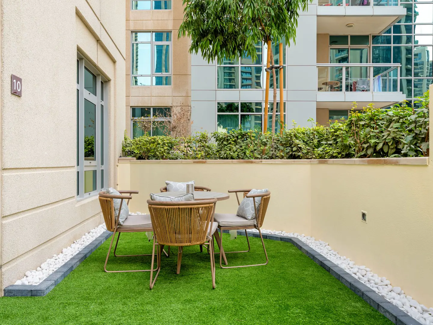 Outdoor patio with a round table and four wicker chairs on green turf, surrounded by white stones and buildings.