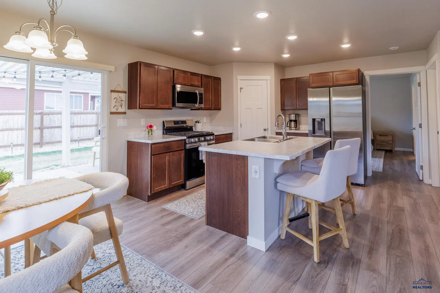 A bright kitchen with wood floors, brown cabinets, and stainless steel appliances. A white island with bar stools faces a dining table near sliding glass doors.