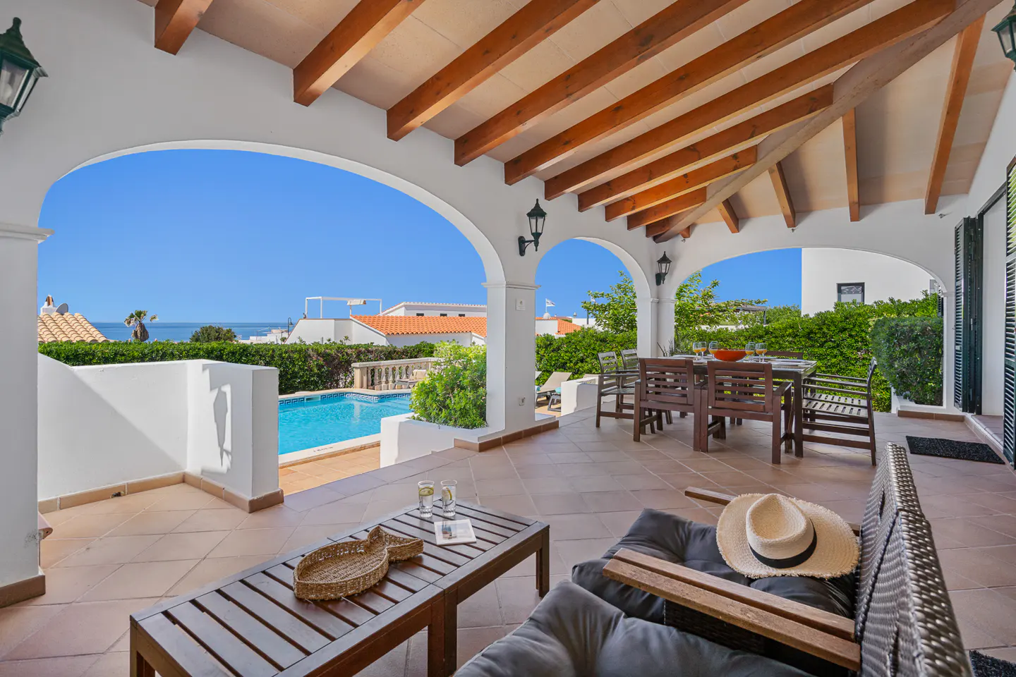 Covered patio with white arches overlooks a pool and ocean. A table and chairs sit to the right, and a lounge chair is in the foreground.