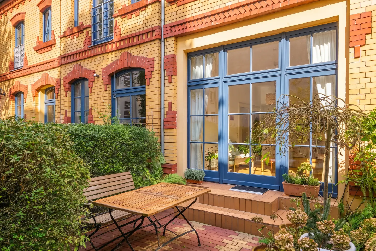 Brick building with blue-framed windows and doors. A wooden table and bench sit on a brick patio with green plants.