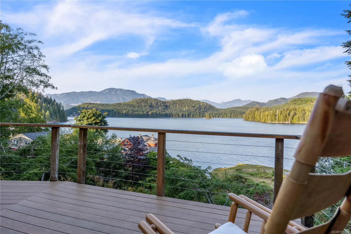 Deck view with a wooden chair overlooking a lake surrounded by green trees and mountains under a blue sky.