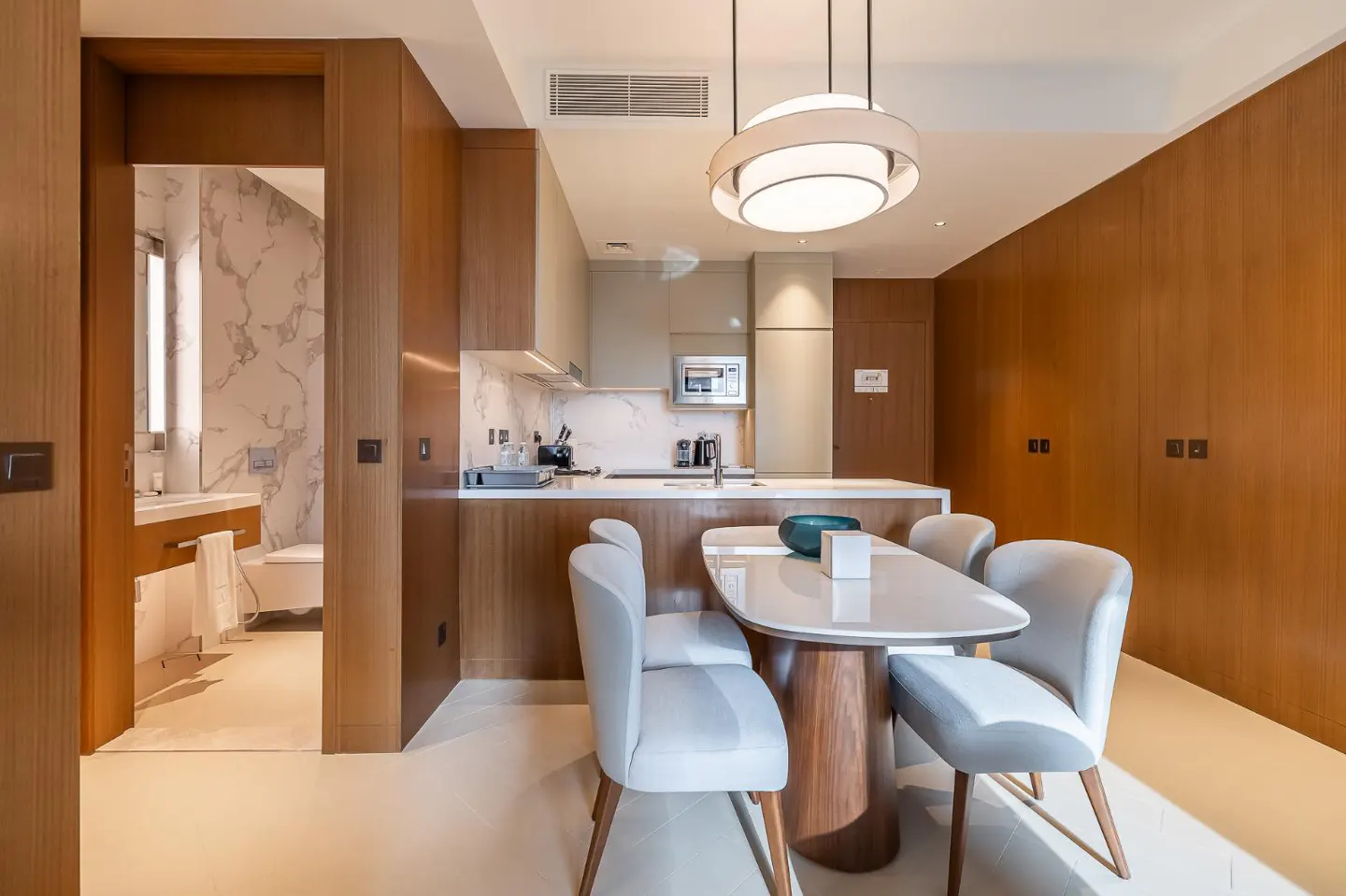 A modern apartment interior with a kitchen, dining area, and bathroom view. The color palette is neutral with wood accents.
