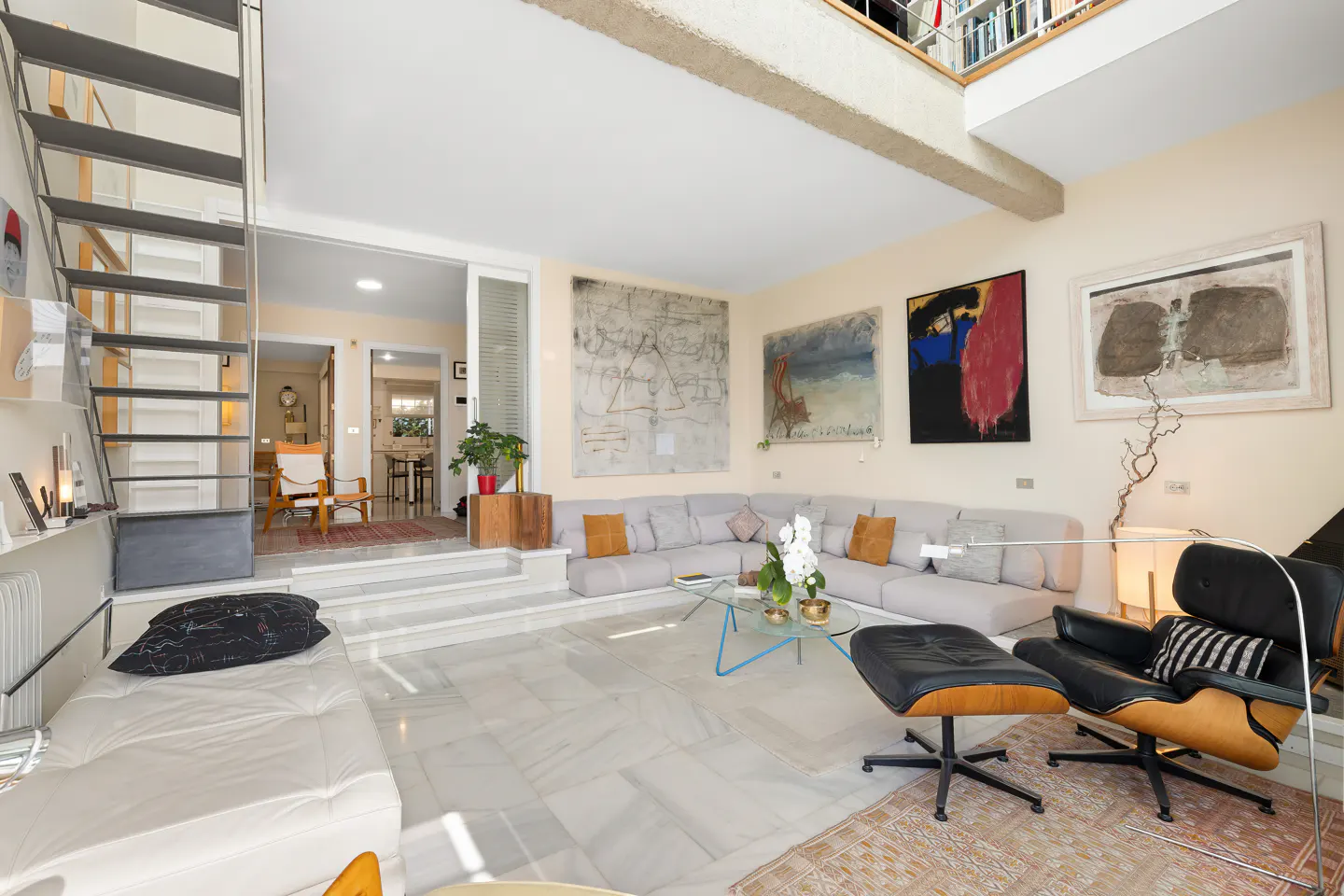 Bright living room with marble floors, a gray sectional sofa, and abstract art. A metal staircase leads to a loft with bookshelves.