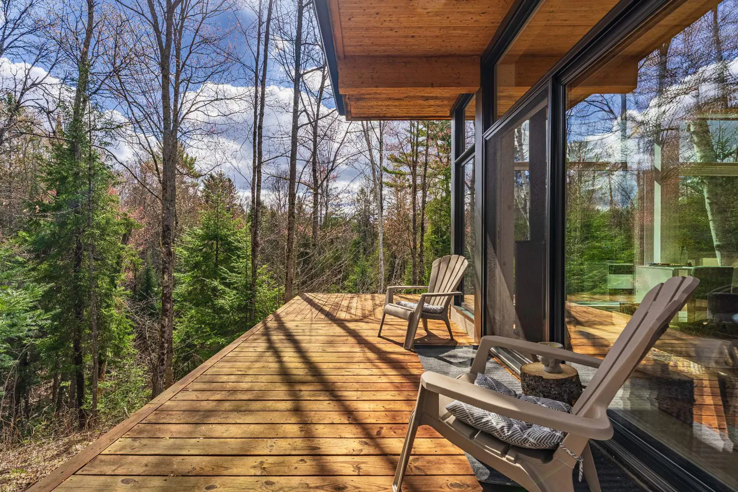 Wooden deck with two Adirondack chairs overlooking a forest. The deck is attached to a modern house with large glass windows.